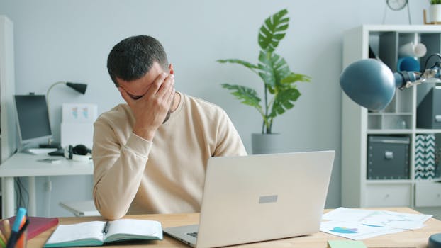 Young man overwhelmed at his desk with laptop, office stress.